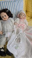 Pair of two vintage dolls seated on a striped pillow and yellow textured fabric, with baby doll accessories in front.