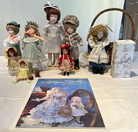 Group photo of all five porcelain dolls standing on doll stands, three smaller dolls, tiny teddy bear, the handled basket, and the doll calendar on a white tablecloth