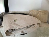 Folded canvas drop cloth with paint splatters and wear marks shown on the left side and an off-white canvas bag with handles and multiple pockets on the right side.