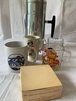 Photo showing the vintage Wear-Ever aluminum coffee pot with black handle, four vintage coffee mugs (Garfield, Tidefest, one with black and white image, one with Tidefest wave design), and a box of filter papers.