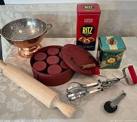 Photo showing copper colander, red tin with seven spices tins inside, Ritz Limited Edition red tin, teal floral tin with lid, wooden rolling pin, hand beater, and metal measuring spoons.