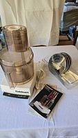 Front view of Cuisinart food processor unit including motor base, work bowl with feed tube, power cord, blade disks, spatula, and instruction manual on a white table.