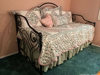Wide view showing full black metal daybed with floral bedding, pillows, and bedskirt against peach wall and green carpet.