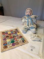 Porcelain doll sitting with knit bonnet, cream dress with blue ribbons, knit footies on feet, pacifier in mouth. To the left, tiny handmade quilt with multicolored fabric yo-yos sewn on cream base. Additional white knit footies visible.