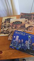 Four Pacific Northwest history books displayed on a wooden table, titles visible including 'Old Tacoma,' 'Alaska Yukon Pacific Exposition,' 'Irish Seattle,' and 'Seattle: The Growth of the City.'