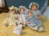 Four porcelain dolls seated on a yellow bedspread, varying sizes from about 15 to 20 inches, dressed in pastel dresses and bonnets. Extra pair of white knit booties in front.