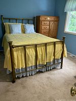 Full size antique brass bed fully made up with yellow chenille bedspread, two blue and white striped pillows, stock blue walls and wood furniture in background