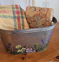 Close-up of the oval galvanized metal bucket painted with colorful floral patterns, containing the two folded tablecloths.
