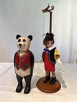 Full view of the hand-carved wooden bear figurine standing next to the string-operated marionette clown doll on a wooden control stand.