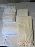 Full view of all linens laid out: folded white twin bedspread with diamond pattern, pillowcases with pink trim and lace, folded curtain panel