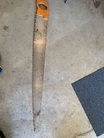 Full view of the entire long saw showing the metal blade with large saw teeth and the wooden handle at one end.