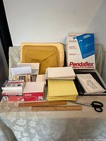 Photo shows variety of office supplies on a cloth surface including Manila envelopes, Pendaflex hanging file folders box, yellow legal pads, white index card boxes, small scissors, ruler