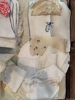 Image showing folded white and beige linens, some embroidered with floral designs in color, lace edges and scalloped hems visible.