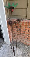 Four metal garden stakes displayed outdoors against brick wall and house siding, showing size and condition.
