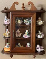 Full view of wooden hanging display case containing 12 porcelain pixie figurines arranged on shelves with some on external side shelves.