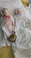 Two porcelain baby dolls lying on lace crocheted blanket. The larger doll has pink ribbon, the smaller blue ribbon. A silver plated cup with spoon and fork is placed near the dolls.