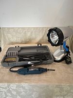 Photo of Ryobi electric detail carver tool in blue with power cord, gray case with accessory carving tips, and Dorcy rechargeable spotlight with charger on table