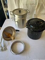 Overview of all items on white cloth, showing aluminum fruit press with wooden pestle, black enamel double pot with lid, and 8-quart aluminum double pot with lid stacked.