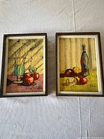 Two wood panel paintings side by side on white cloth background, showing full framed artwork with still life images