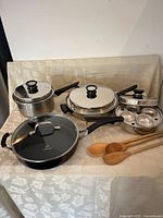 Full view of the set with three stainless steel Queen cookware pieces, one with egg poacher insert, three wooden spoons, and Calphalon pan with glass lid.