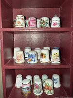 Close-up of three shelves showing varying decorative porcelain thimbles adorned with floral, bird, fruit, and figurative patterns.