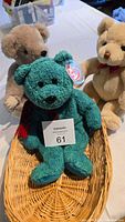 Three bears in a wicker basket on a table: green Beanie Baby Wallace in front, brown jointed bear behind left, cream bear behind right.