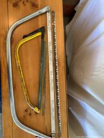Photo showing two bow saw frames, one 36-inch silver metal frame and one 24-inch yellow painted frame, with corresponding saw blades and blade protectors laid flat on a wooden surface.