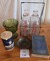 Photo showing all items in lot arranged on wood surface: wire carrier with six glass milk bottles, Mrs. De Graff's Cookbook, Morton Salt container, green glass dish, brass ash tray.