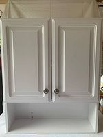Front view of closed white wall-mounted medicine cabinet with two paneled doors and two round knobs, showing bottom open shelf.