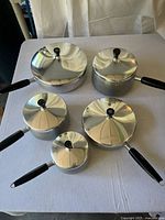 Top-down view of all 5 aluminum WearEver pans with stainless steel lids and black handles, showing assortment of sizes arranged on table.