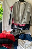 Photo of various clothing items hanging and folded including gray zip-up hoodie, black rain jacket, red button-up shirt, black and white sweaters, denim skorts, white T-shirt, and additional folded clothing.