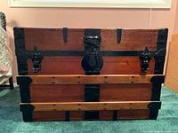 Front view of wooden travel trunk showing stained wood, black metal reinforcements, and hardware including latches and lock mechanism.