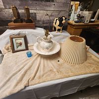 Overview of vintage lot items arranged on cloth including pitcher and bowl lamp, lampshade, framed 3D kittens picture, small cobalt votive holder, and a small floral framed picture.