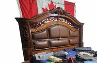 King size bed frame headboard showing carved wood with scroll and floral detailing and upholstered leather panels with nailhead trim, corner light wiring visible.