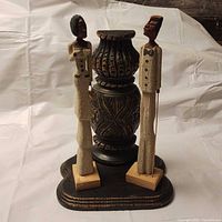 Front view of two tall, slender African figurines in textured formal robes standing on a carved pedestal base with detailed floral motifs and antique-style distressing.