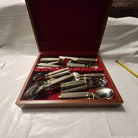 Open wooden storage case with red velvet lining showing neatly arranged flatware including forks, spoons, and knives with ribbed stainless steel handles and gold-tone detailing.