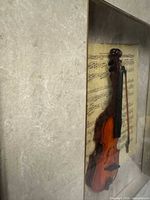 Side-angle photo showing the miniature violin and bow mounted over aged sheet music inside the deep shadowbox frame with glass cover.