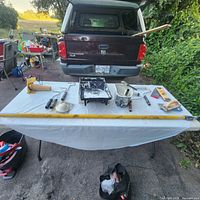 Wide view of an assortment of painting and drywall tools laid out on a table outside with a truck in background.
