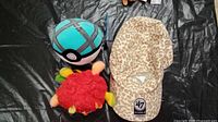 Two plush toys and hat on a black surface: a turquoise and black Pokémon Poké Ball plush, red furry Poppers plush, and leopard print Toronto Blue Jays hat