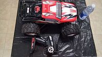 Top view of large red and black RC truck positioned next to a water bottle and black pistol grip remote control on a black plastic surface.