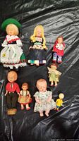 Photo displaying 8 small dolls arranged in two rows on a black surface showing various clothing styles and doll sizes.