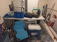 Wide shot showing cooler, folding chair, soft cooler bag, stool, carry bag, rolled mat on and under table