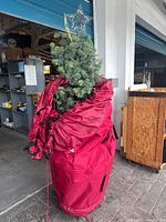 Artificial green Christmas tree with silver star topper partially inside a red fabric storage bag.
