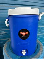 Front view showing blue cylindrical cooler with white lid, handle and white spigot at base, Coleman brand molded on the cooler body with a black-red D-DEY sticker.
