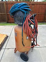 Front angle showing orange tank with motor housing and coiled blue and red air hoses wrapped around the unit. Two black wheels visible on bottom for mobility.