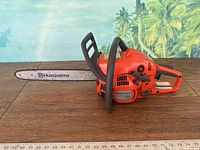 Full side view of orange Husqvarna 240 chainsaw on wooden surface with tropical background.