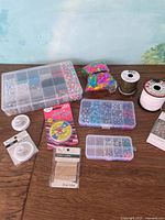 Full shot of lot showing two plastic bead containers, colorful clips wrapped in rubber band, packaged thread and wire spools, and jewelry accessories on wooden surface.