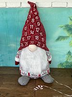 Front full view of large plush gnome advent calendar showing red hat with advent numbers, white beard, and gray feet, with one candy cane placed.