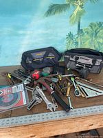 Various hand tools laid out including wrenches, saw, pliers, clamps, measuring tape, hammer, screwdriver, square, and metal ruler, along with two branded tool bags.