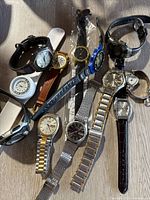 Lot of 11 preowned wristwatches on a wood surface, showing variety of band types including metal links, leather, and plastic, in fair to heavily used condition.